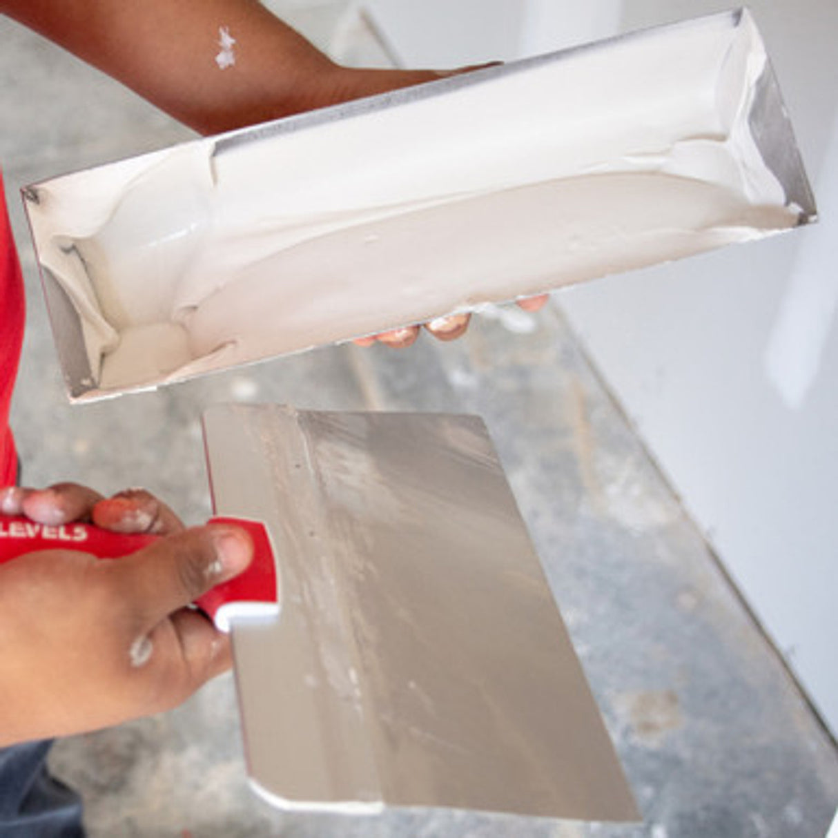 Person holding a Level 5 taping knife and a mud pan full of compound at a job site