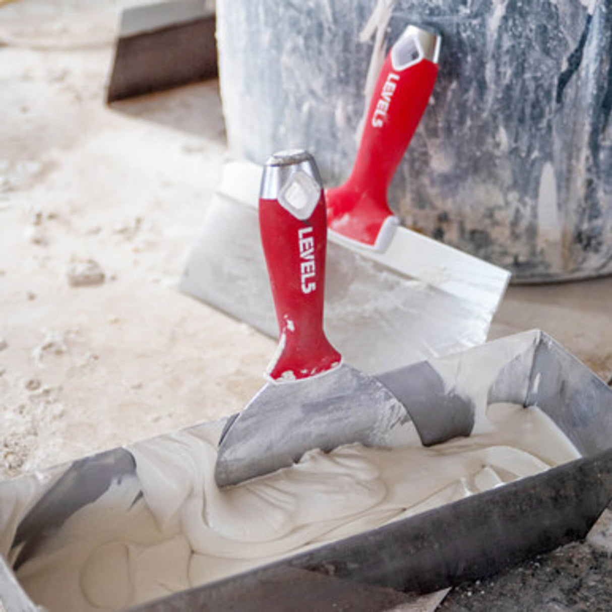 Level 5 joint knife in a mud pan filled with compound, with a Level 5 taping knife resting on a bucket in the background on a jobsite floor